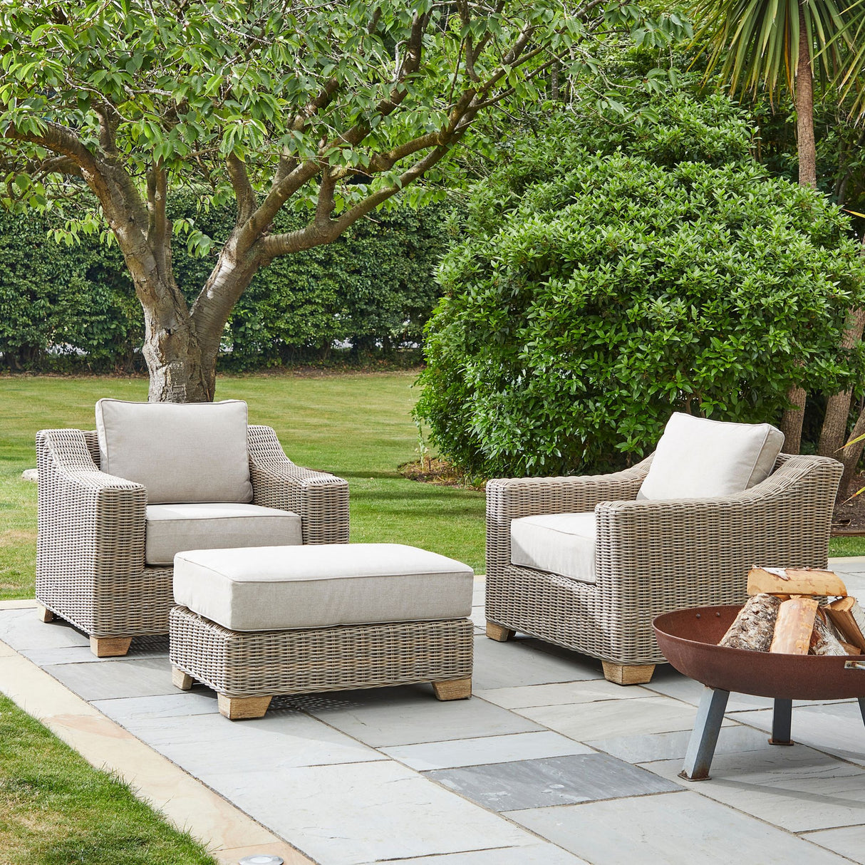Hill Interiors beige woven outdoor foot stool with cushioned top, set on a stone patio between two matching armchairs beside a fire pit and garden.
