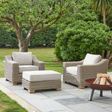 Hill Interiors beige woven outdoor foot stool with cushioned top, set on a stone patio between two matching armchairs beside a fire pit and garden.