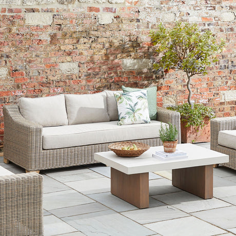 Hill Interiors Rattan Three-Seater Garden Sofa in beige with cushions and a palm-print pillow, set on a patio beside a wooden coffee table against a brick wall.