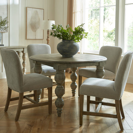 Hill Interiors Grey Wooden Dining Chairs with nailhead trim and neutral upholstery, set around a round wooden table in a sunlit dining room.