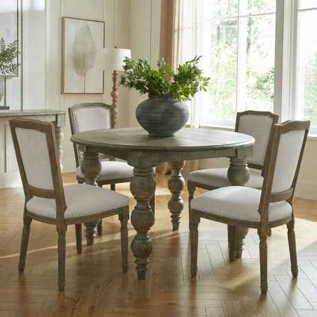 Hill Interiors grey wooden dining chair with upholstered back and seat, nailhead trim, and turned legs, set around a round table in a bright dining room.