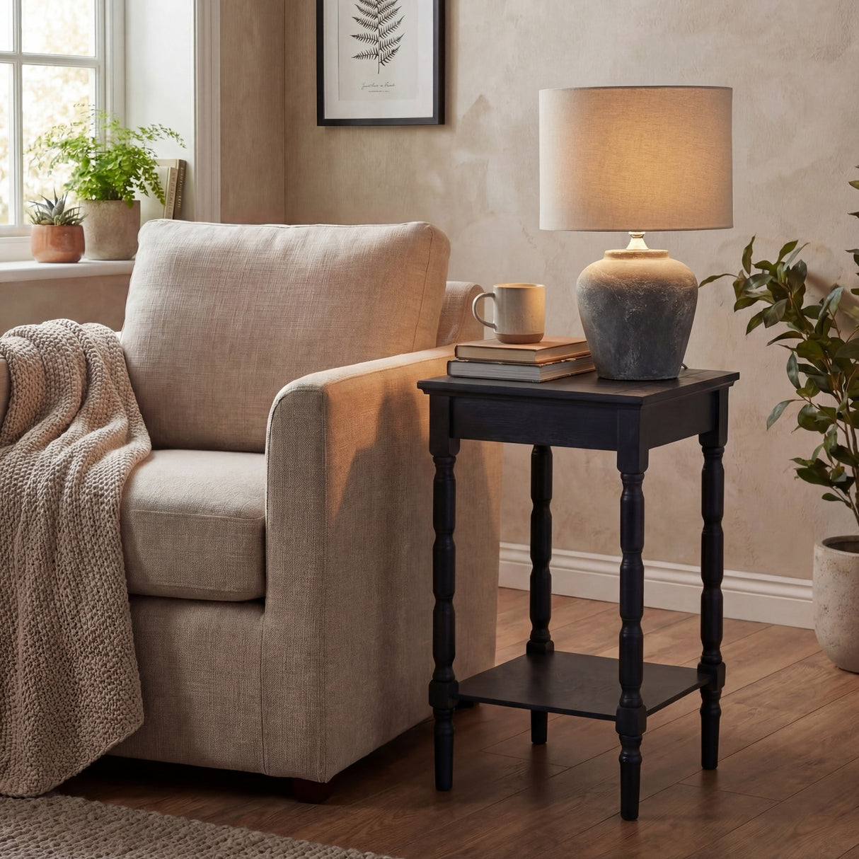 Hill Interiors Ferranock grey ceramic table lamp with rounded base and natural linen drum shade, lit on a black side table in a neutral living room.