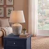 Hill Interiors Merenza antique white ceramic table lamp with rounded tapered base and natural linen shade, styled on a navy side table by a window.