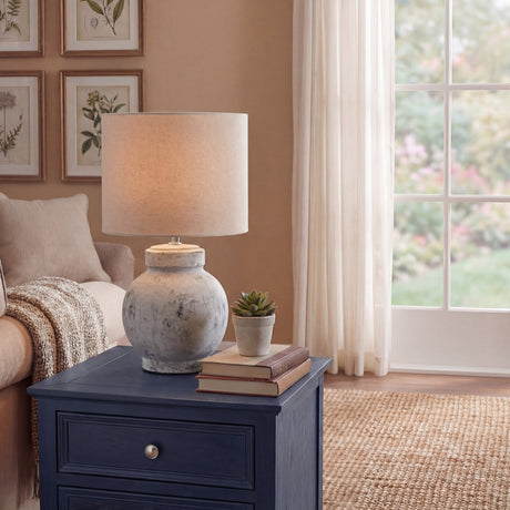 Hill Interiors Merenza squat ceramic table lamp with antique white glazed base and natural linen drum shade, styled on a navy side table by a window.
