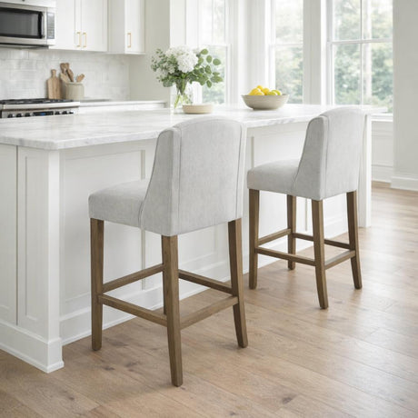 Pair of Hill Interiors Braxdon grey wooden bar stools with curved upholstered backs at a white marble kitchen island with wood floor.