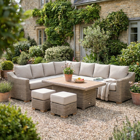 Hill Interiors Large Outdoor Corner Set with beige cushions, wicker-effect metal frame, timber-topped riser table and two stools on a gravel garden patio.
