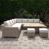 Hill Interiors large beige outdoor corner sofa set with cushioned L-shaped seating, timber-topped riser table and two matching stools on a patio.