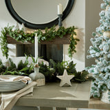 Hill Interiors small white ceramic standing star decoration on a wood table with pine garland, candle, and plates near a flocked Christmas tree and fireplace.