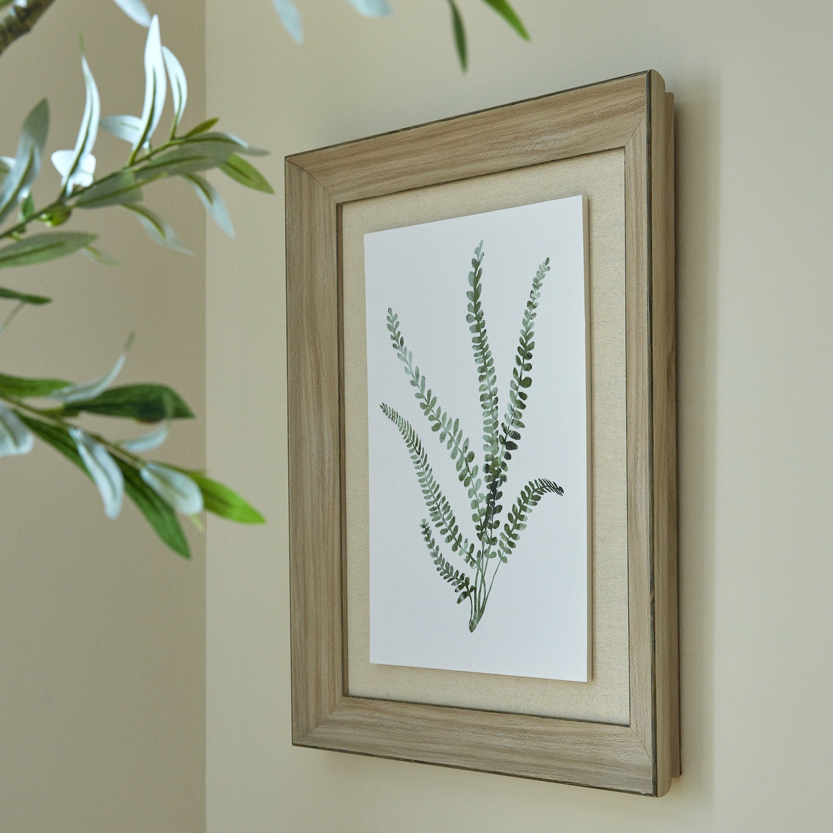 Hill Interiors Delicate Fern Print in a washed pale brown wood frame, angled on a neutral wall beside soft green foliage in a living space.