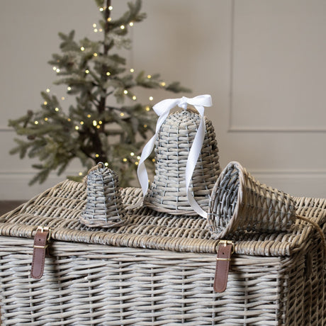Hill Interiors large wicker bell decoration in brown with white ribbon, displayed with a smaller bell on a woven basket beside a lit Christmas tree.