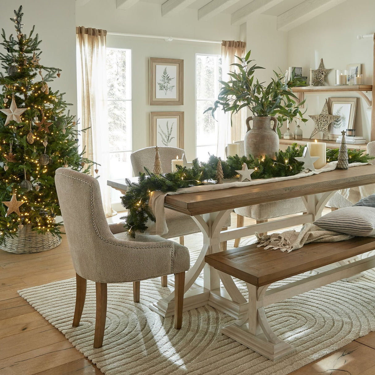 Festive dining room styled with Hill Interiors wicker baubles on a lit Christmas tree, beside a wooden table with garland, candles and neutral chairs.