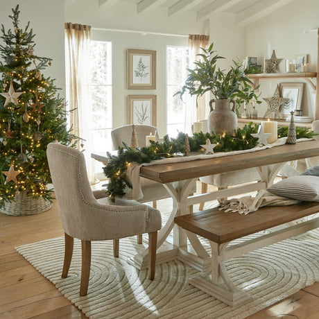 Festive dining room styled with Hill Interiors wicker baubles on a lit Christmas tree, beside a wooden table with garland, candles and neutral chairs.