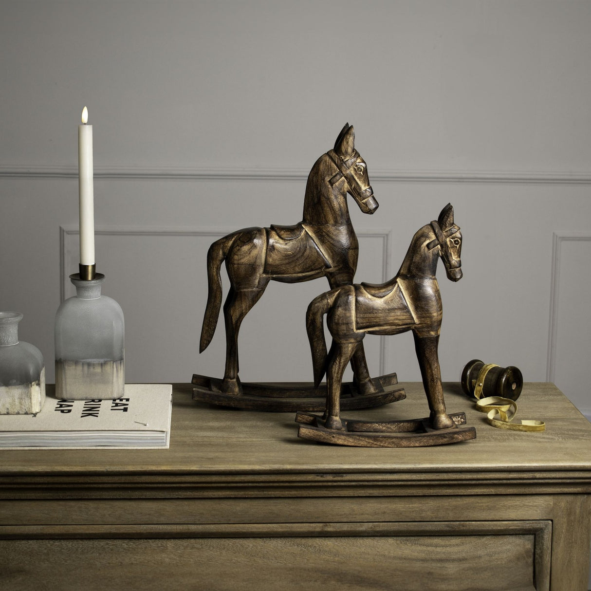 Hill Interiors wooden rocking horse decorations in beige, shown as two carved horses on curved rocker bases styled on a wooden sideboard with a candle.