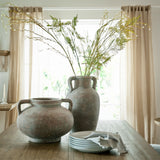 Hill Interiors Large Ceramic Amphora Vase in brown with tall fern stems, beside a smaller handled pot and stacked plates on a wooden dining table.