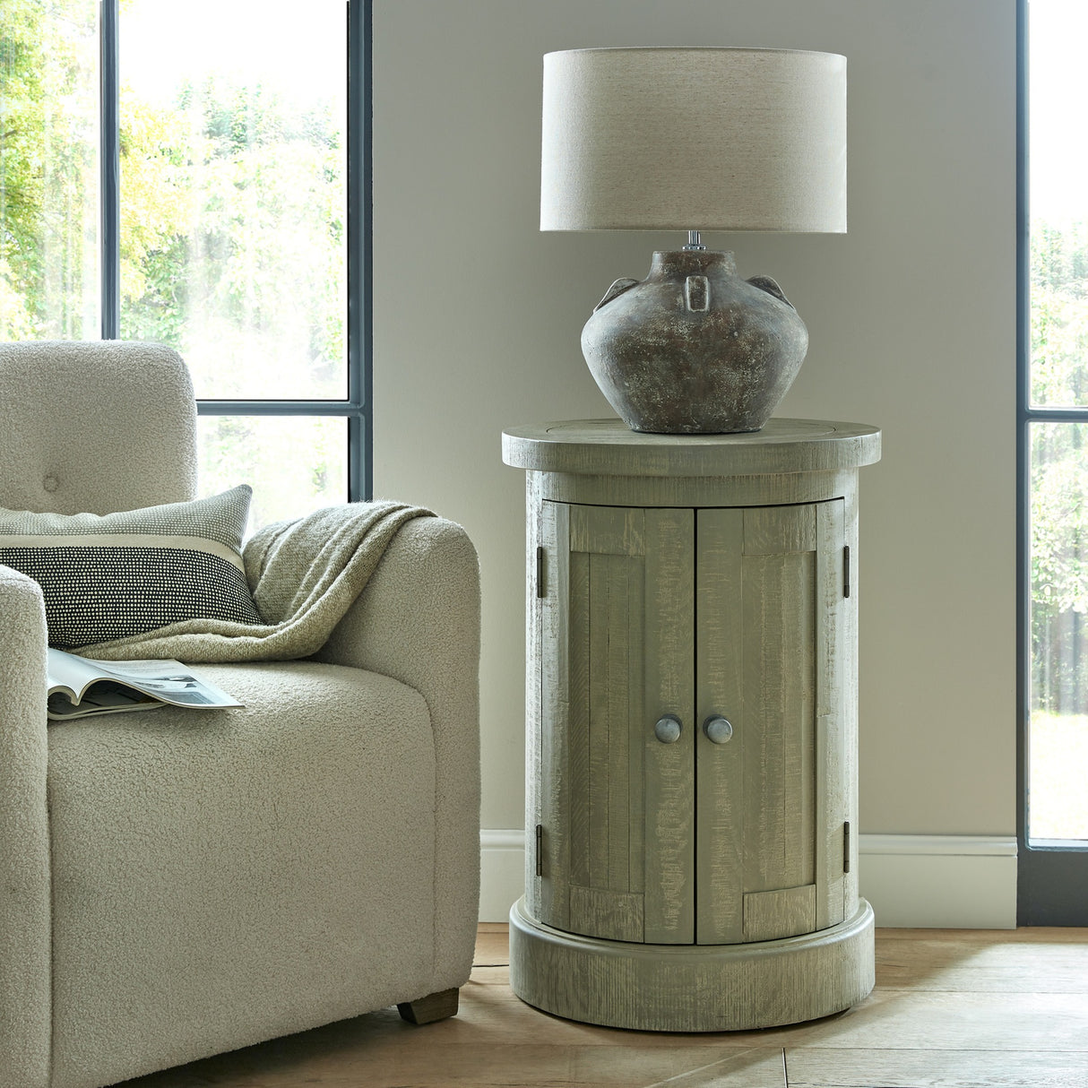 Hill Interiors Lumbriden Lekanis ceramic table lamp with brown urn-shaped base and drum shade on a round wooden cabinet in a bright living room.