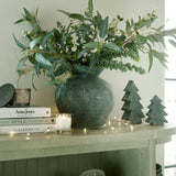 Hill Interiors ceramic medium Christmas trees in grey displayed on a console table with a foliage-filled vase, candle, books and fairy lights.