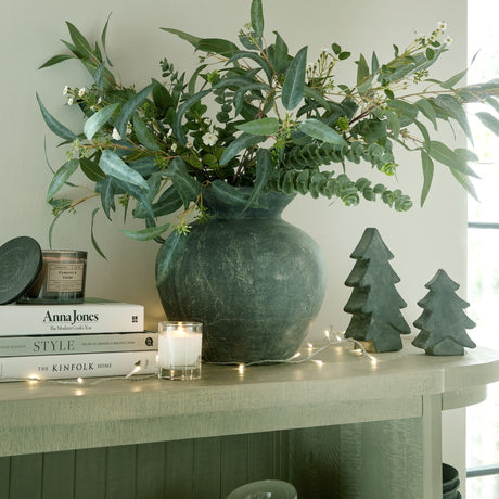Hill Interiors small grey ceramic Christmas tree styled on a console with a larger tree, foliage-filled vase, books, candle and fairy lights.