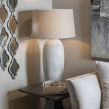 Hill Interiors Elvessa beige ceramic table lamp with ribbed urn-shaped base and drum shade, lit on a dark side table beside a mirror and artwork.