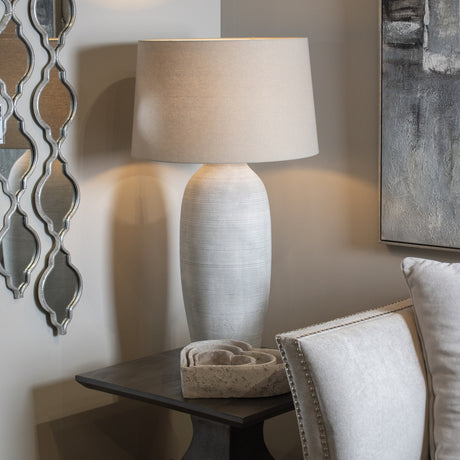 Hill Interiors Elvessa beige ceramic table lamp with ribbed urn-shaped base and drum shade, lit on a dark side table beside a mirror and artwork.