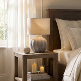 Hill Interiors Merenza fluted ceramic table lamp in antique white with linen drum shade, styled on a wooden bedside table beside a bed and window.