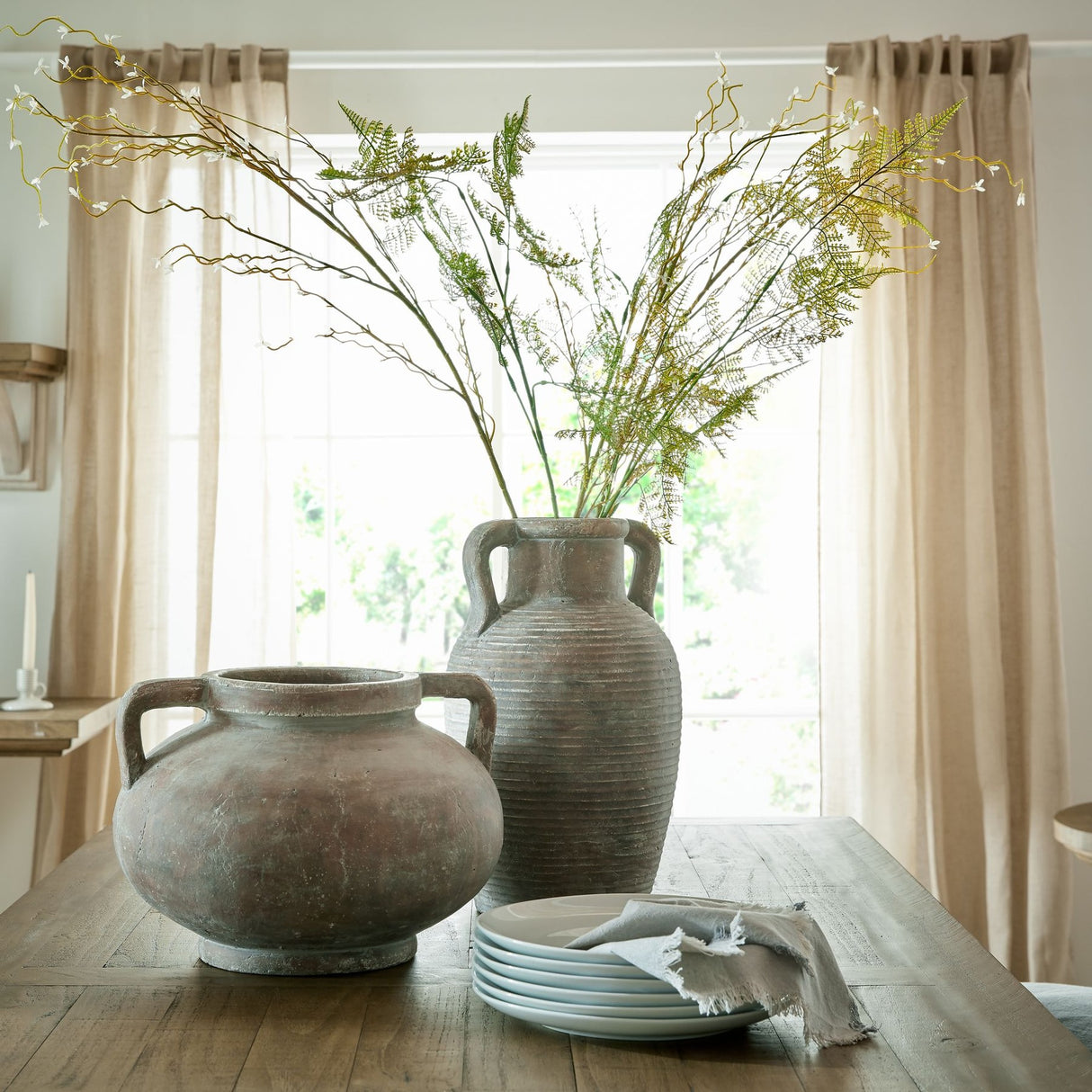 Hill Interiors brown ceramic pelike pot vase with handles holding tall fern and blossom stems, beside a rounded handled pot on a wooden dining table.