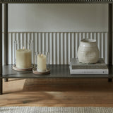 Hill Interiors ceramic hurricane lanterns with lit pillar candles beside stacked books and a textured cream jug on a shelf against panelled walls.