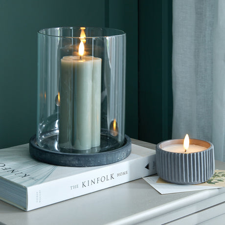 Hill Interiors Large Hurricane Lantern in grey with a lit pillar candle inside the clear glass cylinder on a ceramic base, styled on a book beside a tealight.