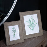 Hill Interiors Woven Photo Frame in grey with textured wooden surround, shown in two sizes displaying botanical prints on a wooden dresser beside a mirror.