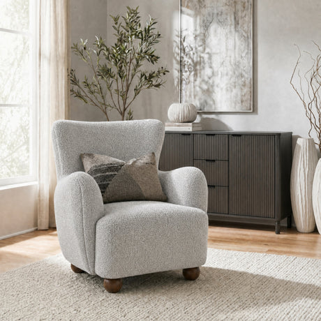Hill Interiors large beige bouclé accent chair with wooden bun feet and patterned cushion, styled in a neutral living room beside a dark sideboard.