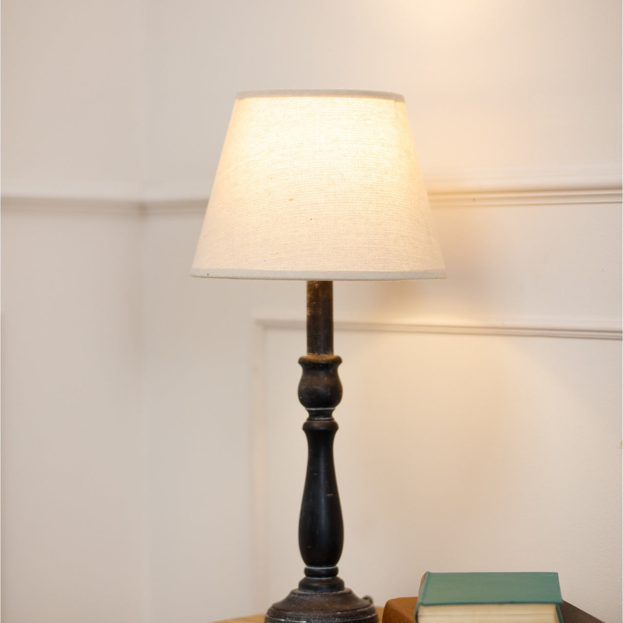 Hill Interiors Column Table Lamp with turned grey wood base and natural linen tapered shade, lit on a side table beside books against a pale wall.