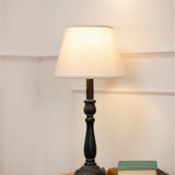 Hill Interiors Column Table Lamp with turned grey wood base and natural linen tapered shade, lit on a side table beside books against a pale wall.