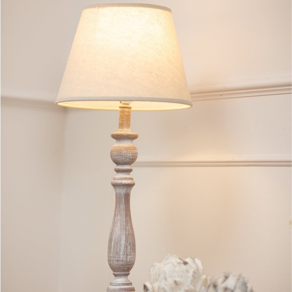 Hill Interiors Column Table Lamp with turned white wooden base and natural linen shade, illuminated on a console beside a pale decorative accent.