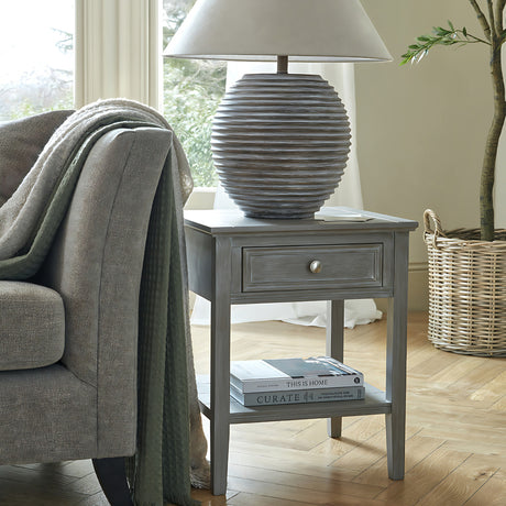 Hill Interiors Wooden One-Drawer Side Table in grey beside an armchair, holding a ribbed ceramic lamp, with books stacked on the lower shelf.