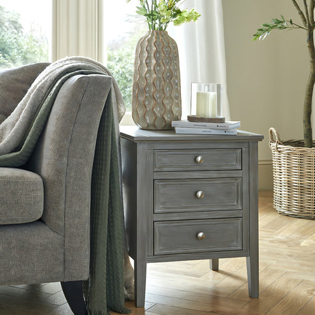 Hill Interiors Wooden Three-Drawer Bedside Table in grey with round metal knobs, styled beside a woven armchair with vase, candle and books on top.