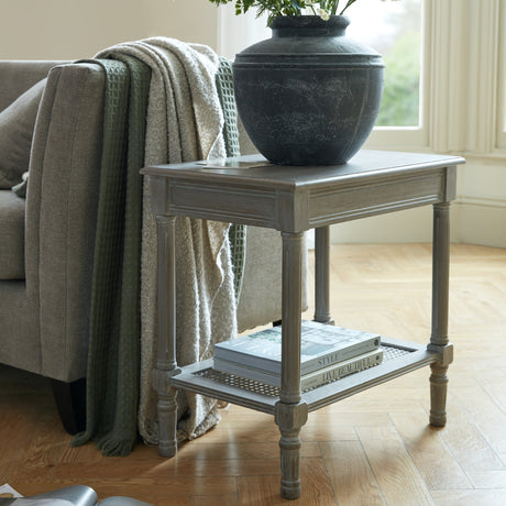 Hill Interiors Rattan Side Table in grey with turned wood legs and woven lower shelf holding books, styled beside a sofa with a dark vase on top.