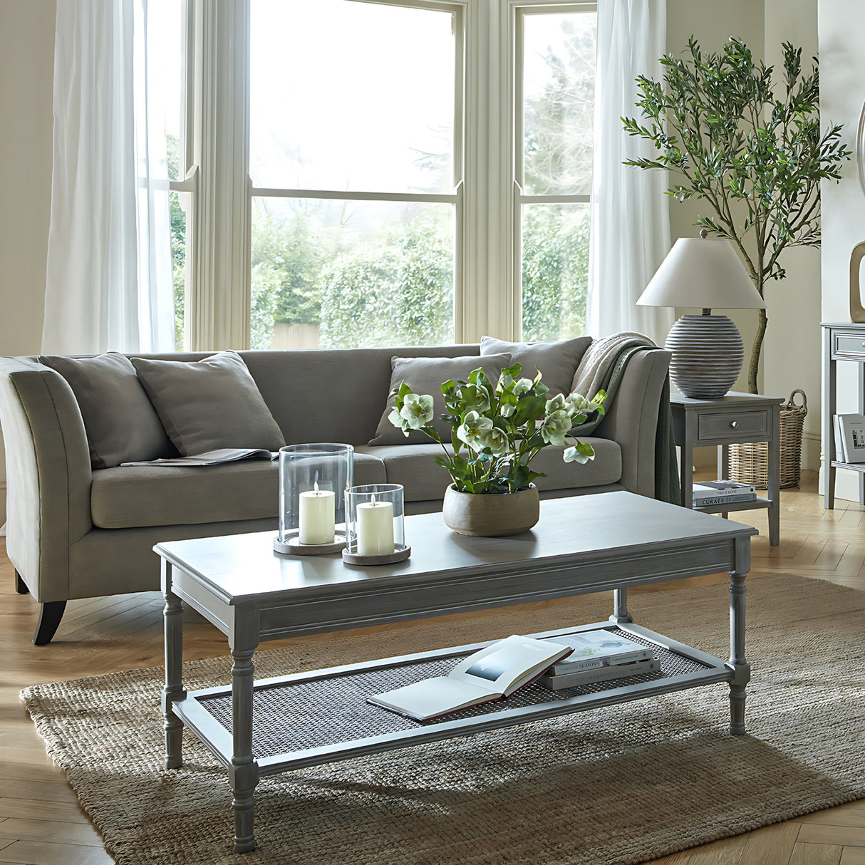 Hill Interiors Rattan Coffee Table in brown with woven lower shelf, styled with candles and flowers in a bright living room with grey sofa.