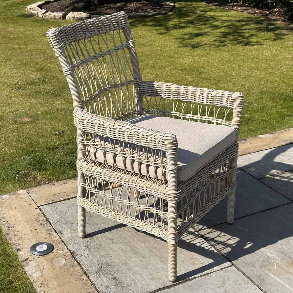 Hill Interiors beige metal outdoor dining chair with woven rattan-effect frame and cushioned seat, set on a garden patio beside a lawn.