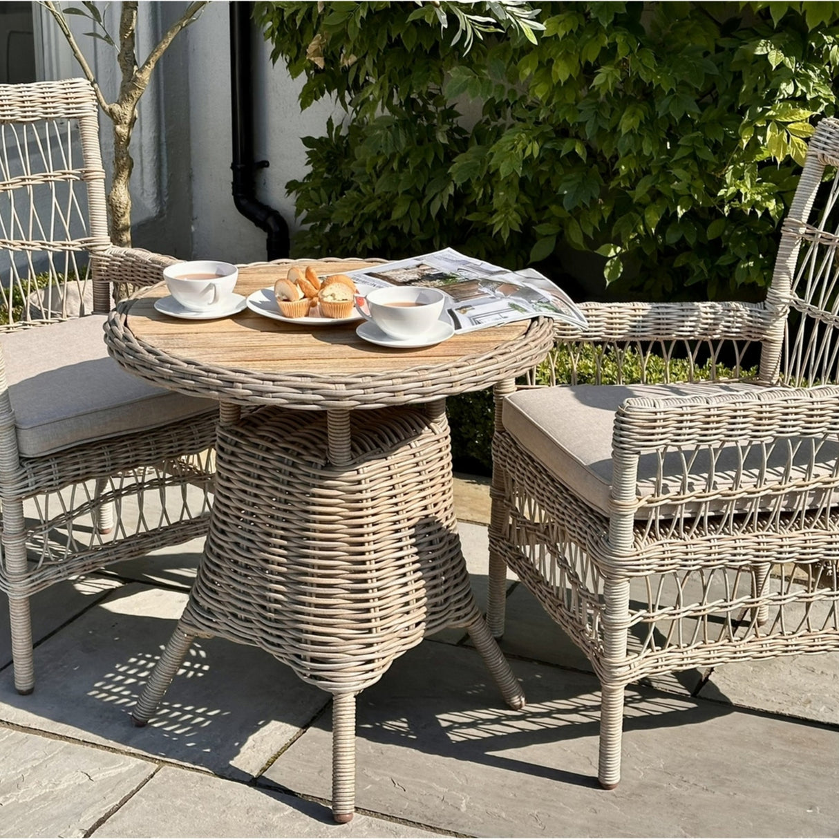 Hill Interiors beige metal outdoor dining chairs with cushions arranged around a round woven bistro table set with tea and pastries on a sunny patio.