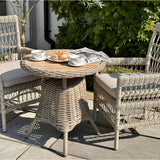 Hill Interiors beige metal outdoor dining chairs with cushions arranged around a round woven bistro table set with tea and pastries on a sunny patio.