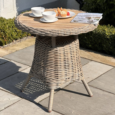 Hill Interiors beige bistro table with round wood top and woven rattan-effect base on a patio, styled with teacups, pastries and a magazine.
