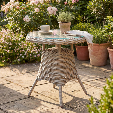 Hill Interiors beige woven rattan-effect bistro table with round glass top holding a teacup, potted herb and linen napkin on a sunlit garden patio.