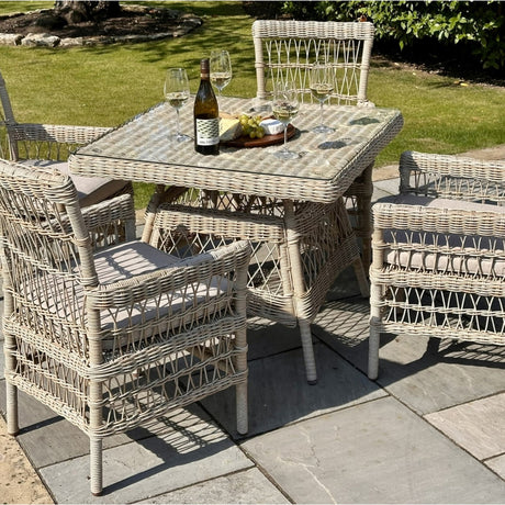 Hill Interiors Outdoor 4 Seater Dining Set with beige woven rattan chairs and square glass-top table on a garden patio, set with wine and cheese.