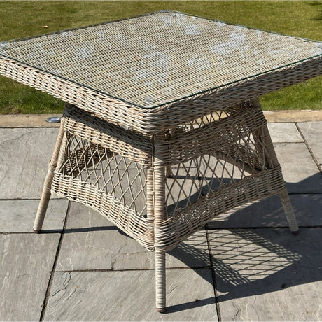 Hill Interiors Outdoor 4 Seater Dining Set with a square beige woven rattan table and clear glass top, shown on a stone patio beside a lawn.