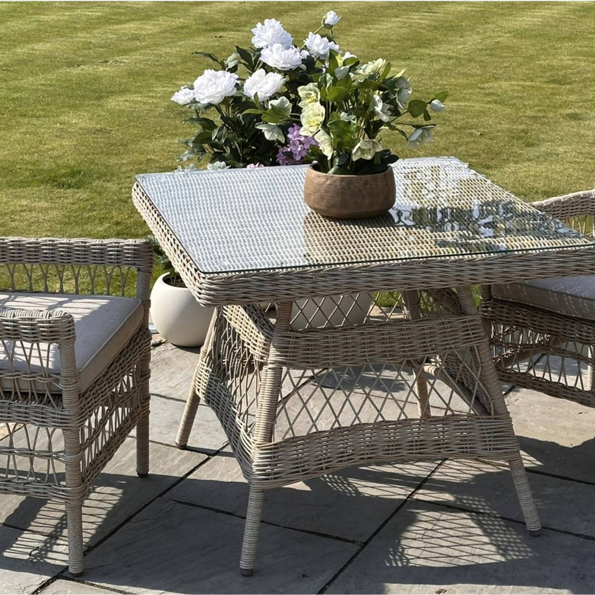 Hill Interiors Outdoor 4 Seater Dining Set with beige woven rattan chairs and square glass-top table on a garden patio beside a lawn.