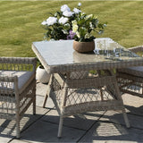 Hill Interiors Outdoor 4 Seater Dining Set with beige woven rattan chairs and square glass-top table on a garden patio beside a lawn.