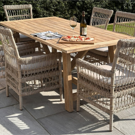 Hill Interiors Large Outdoor Wooden Dining Table in brown, set on a patio with woven rattan armchairs, styled with wine and pizza in a garden.