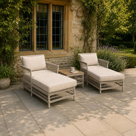 Hill Interiors Rattan Garden Sun Lounger Set in beige, a pair of woven reclining loungers with cream cushions on a stone patio beside a side table.