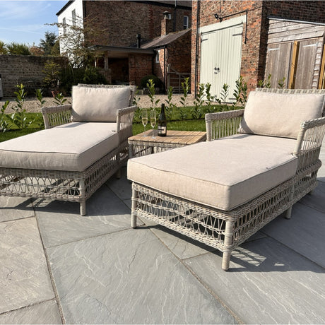 Hill Interiors Rattan Garden Sun Lounger Set in beige, a pair of woven reclining loungers with cushions on a stone patio beside a brick house.