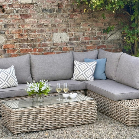 Hill Interiors Outdoor Large Corner Set in beige with woven rattan base, grey cushions, patterned pillows and glass-topped coffee table against a brick wall.