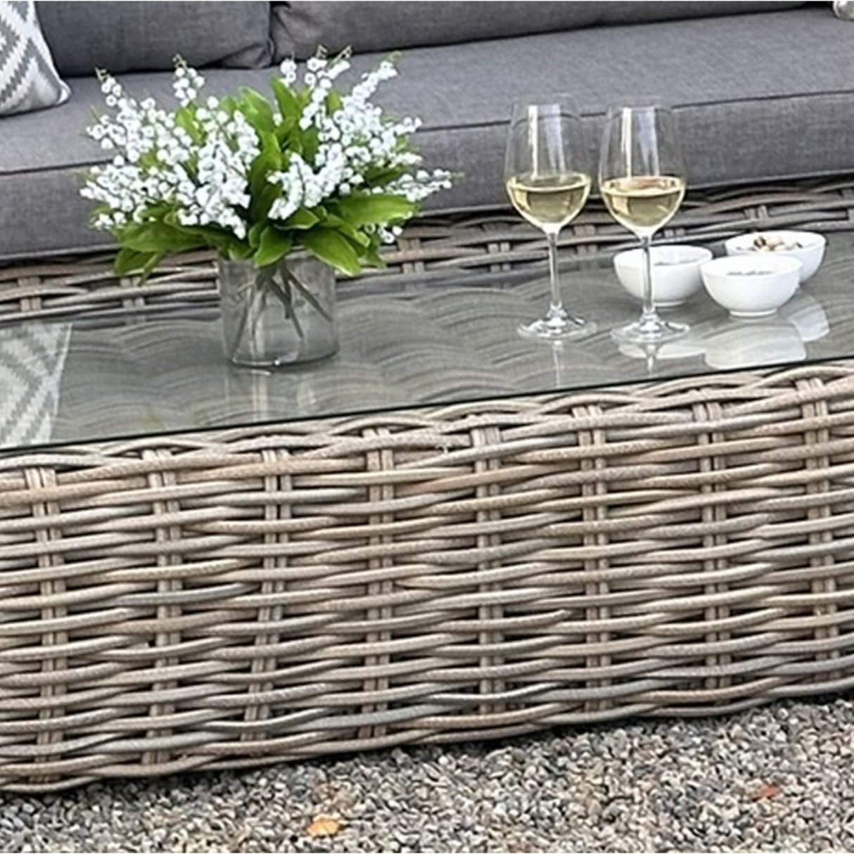 Hill Interiors Outdoor Large Corner Set in beige with woven rattan base, glass top holding two white wine glasses, small bowls and a vase of white flowers.